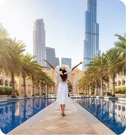 Woman in a white dress and hat stands with arms raised, facing palm trees and skyscrapers, with a reflecting pool in front.
