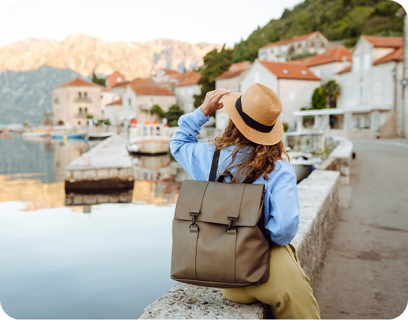 Person wearing a hat and backpack sits by a waterfront, looking at distant hills, with quaint buildings lining the water's edge.