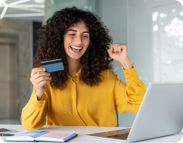 A woman in a yellow shirt celebrates while holding a credit card and looking at a laptop.